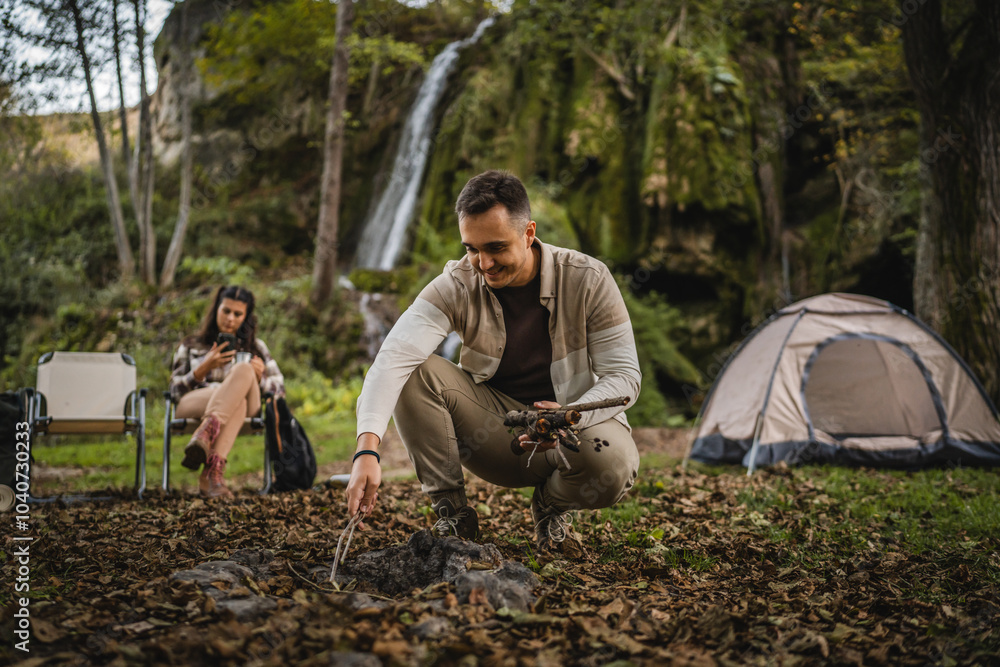 Adult man camper sets up a campfire in front a tent and waterfall Adult man camper sets up a campfire in front a tent and waterfall
