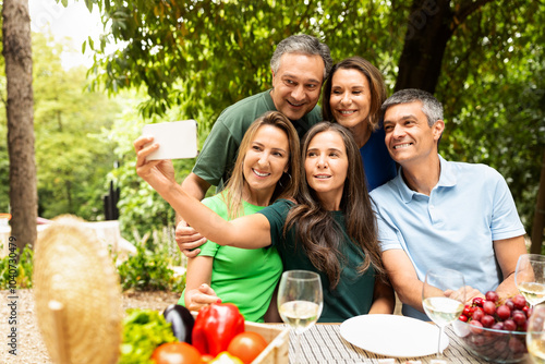 A group of five friends gathers around a table covered with fresh fruits and drinks. They smile joyfully as they take a selfie together in a vibrant, green park during a sunny day. photo