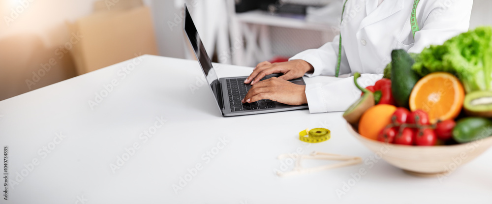 Fototapeta premium Cropped of fresh fruits and vegetables bowl next to working online african doctor, dietician using laptop