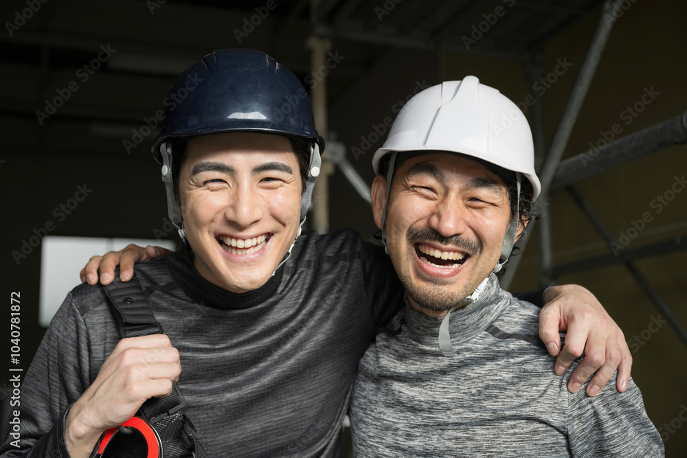現場作業員(鳶職や屋外の安全帯ハーネスをつけた）の肩を組むガテン系仲良し2人の見上げるカメラ目線の笑顔　転職や採用のイメージ