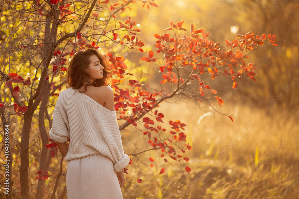 A young brunette woman in a cozy off-shoulder sweater poses near a tree with vibrant autumn leaves, bathed in warm golden light. Perfect for fall fashion, lifestyle, or seasonal promotions. A young brunette woman in a cozy off-shoulder sweater poses near a tree with vibrant autumn leaves, bathed in warm golden light. Perfect for fall fashion, lifestyle, or seasonal promotions.