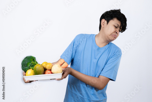 Young excited Indonesian man standing carrying tray of healthy vegetables and fruits with his hand pointing, healthy lifestyle concept, healthy food campaign, isolated on white background. photo