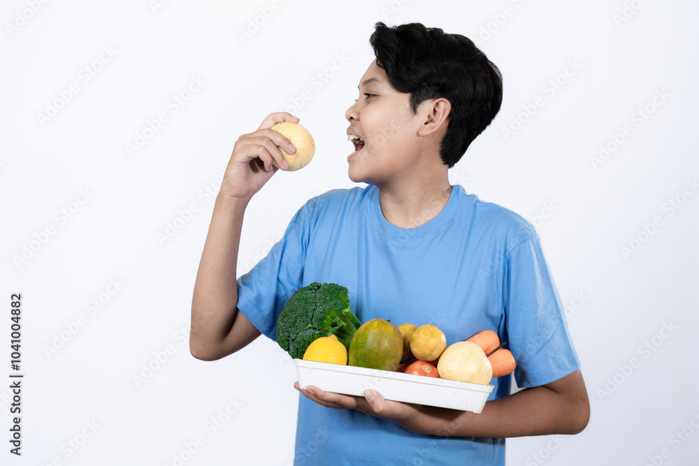 Obraz premium Young excited Indonesian man standing carrying tray of healthy vegetables and fruits with his hand pointing, healthy lifestyle concept, healthy food campaign, isolated on white background.