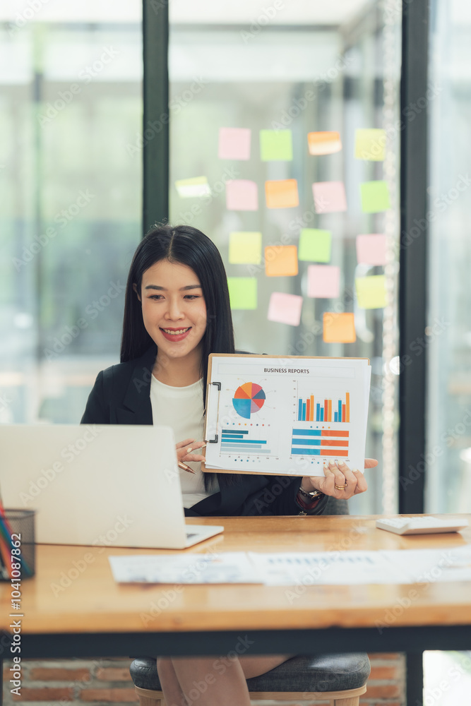 Confident Businesswoman Presenting Data: A young, smiling Asian businesswoman confidently presents business charts and graphs during a video conference, showcasing professionalism and success. Confident Businesswoman Presenting Data: A young, smiling Asian businesswoman confidently presents business charts and graphs during a video conference, showcasing professionalism and success.