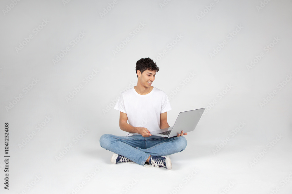 Chico sentado en el suelo con vaqueros y camiseta blanca, mirando un portátil. Concepto de trabajo remoto, estudio online y venta digital. Chico sentado en el suelo con vaqueros y camiseta blanca, mirando un portátil. Concepto de trabajo remoto, estudio online y venta digital.