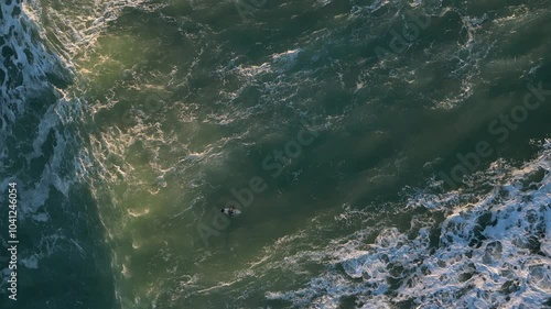 Top-down drone view of a surfer sitting on their board during sunset, surrounded by calm ocean waters and warm evening light.