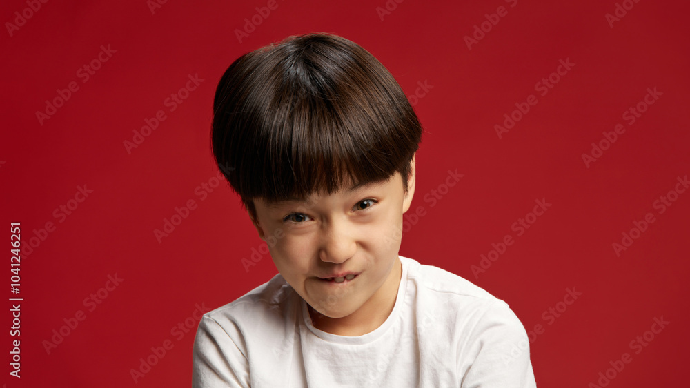 Child, little boy with playful smirk, biting his lip as if planning something mischievous. against bold red studio background. Concept of facial-expressions, positive and negative emotions. Ad Child, little boy with playful smirk, biting his lip as if planning something mischievous. against bold red studio background. Concept of facial-expressions, positive and negative emotions. Ad
