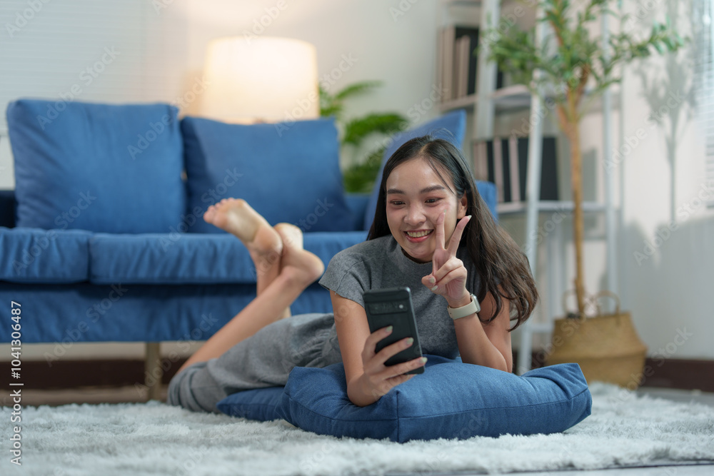 Happy young attractive Asian woman lying on carpeted area in living room using mobile phone connected to the internet to play games. online shopping View social media, online chat, technology conce Happy young attractive Asian woman lying on carpeted area in living room using mobile phone connected to the internet to play games. online shopping View social media, online chat, technology conce
