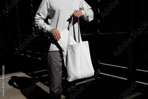 Man pulling a book from the tote bag mockup template in an urban outdoor environment with sharp sunlit shadows. Isolated surface to place your design. 