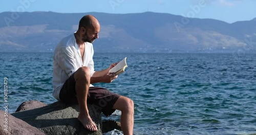 A middle-aged man sitting on the rocks by the seaside, reading a book on a summer day