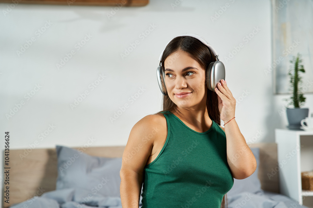 Naklejka premium A young brunette woman with short stature listens to music in her cozy bedroom, reveling in her peaceful time at home.