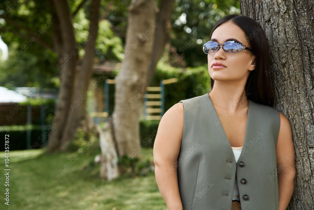 A young brunette woman with a confident expression relaxes by a tree, soaking up the sun in a vibrant outdoor setting. A young brunette woman with a confident expression relaxes by a tree, soaking up the sun in a vibrant outdoor setting.