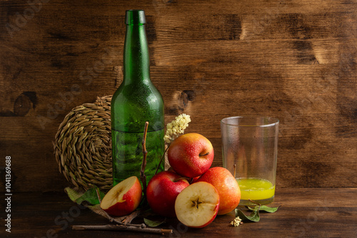 Close-up of bottle and glass with natural cider on table and wooden background with apples, leaves in horizontal with copy space