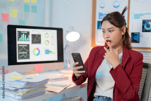 Young asian businesswoman working late in the office looks shocked reading her phone, reflecting stress and pressure of corporate life