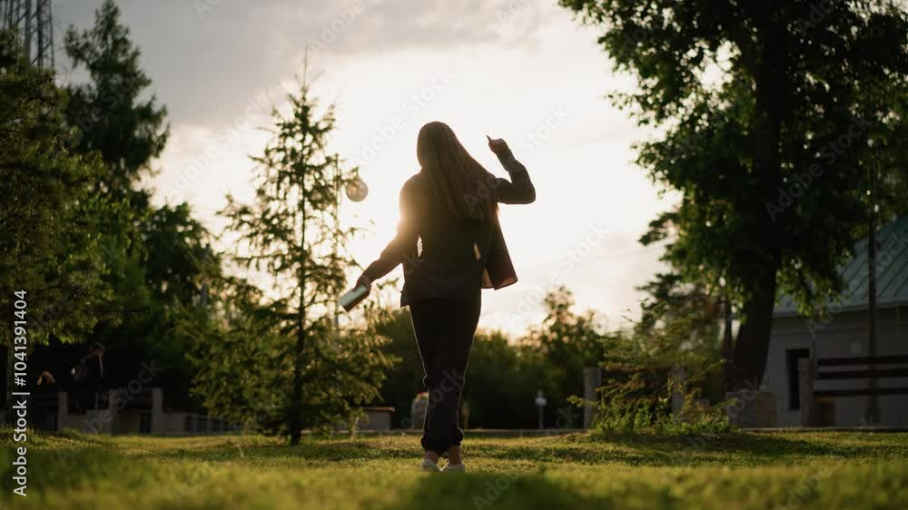 Back view of Lady joyfully dancing outdoors in grassy field with book in hand as sunlight creates radiant atmosphere around her, with a mix of greenery, trees, and building in background Back view of Lady joyfully dancing outdoors in grassy field with book in hand as sunlight creates radiant atmosphere around her, with a mix of greenery, trees, and building in background