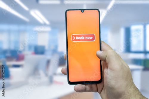 Close-up of the “Book Now” message on the smartphone screen. Online booking concept. Office background. Early booking message. 