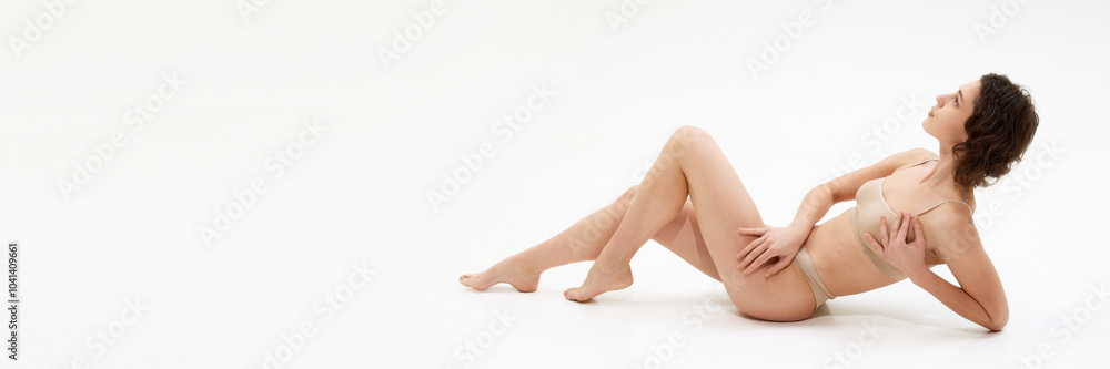 Obraz premium Banner, Young woman sitting in beige lingerie on floor in relaxed posture against white studio background with negative space to insert text. Concept of spa procedure, self-care treatments, self-love.