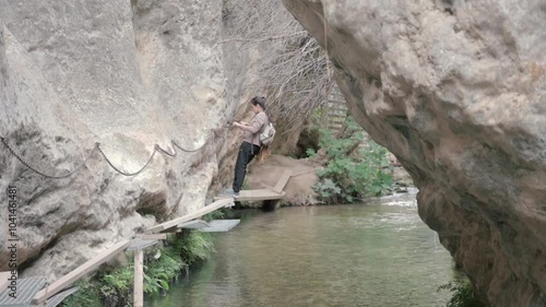 Hiker going across a rocky riverbed, holding onto a chain for support while walking on narrow, precarious wooden walkways photo