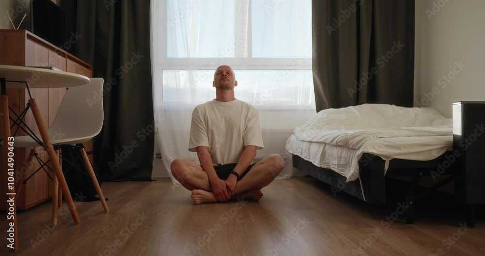 A freelance man is sitting on the floor in his bright apartment against the background of a window. A man does breathing exercises to calm down and distract himself from his work. High quality 4k A freelance man is sitting on the floor in his bright apartment against the background of a window. A man does breathing exercises to calm down and distract himself from his work. High quality 4k