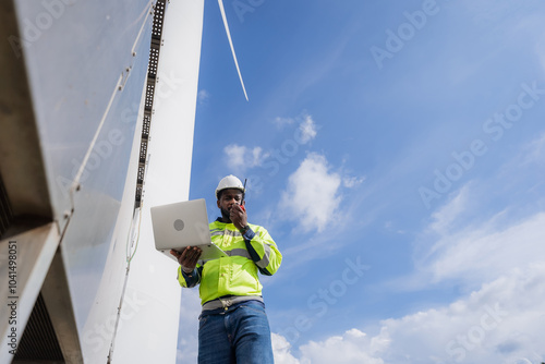 A man in a yellow safety vest is talking on a cell phone while looking at a laptop. electrical, engineer, environment, energy, clean, sustainable, wind turbine, nature