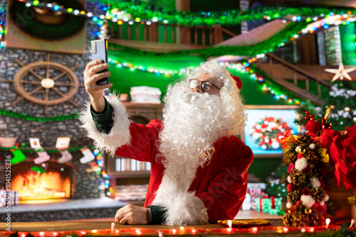 Old Father Christmas takes photos of himself in the festive red suit, having fun during the winter holidays to spread the x mas cheer. Santa taking pictures on his smartphone, modern tradition. photo