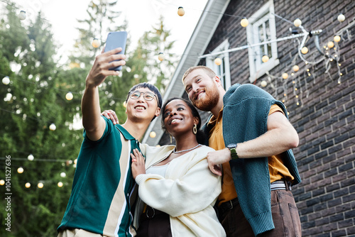 Low angle portrait of multiethnic group of adult friends taking selfie together outdoors in house yard decorated with fairy lights copy space photo