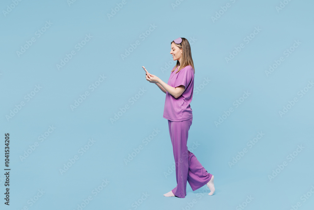 Full body side view calm young woman wear purple pyjamas jam sleep eye mask rest relax at home hold in hand use mobile cell phone isolated on plain blue cyan background. Good mood night nap concept. Full body side view calm young woman wear purple pyjamas jam sleep eye mask rest relax at home hold in hand use mobile cell phone isolated on plain blue cyan background. Good mood night nap concept.