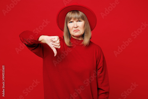 Elderly sad displeased dissatisfied blonde woman 50s years old wear sweater hat casual clothes showing thumb down dislike gesture isolated on plain red background studio portrait. Lifestyle concept. photo