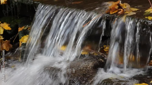 Waterfall cascading down rocky terrain, with yellow leaves drifting on the water and foam forming at the base. A calm, seasonal moment in the heart of nature. photo