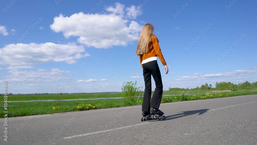 A girl with long hair on roller skates rides along the road past a green field and blue sky. A girl with long hair on roller skates rides along the road past a green field and blue sky.