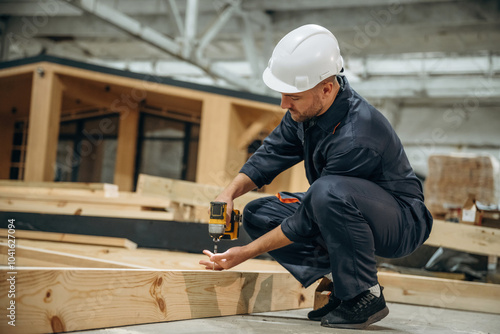 Small drill in hands. Male builder is at construction of wooden house