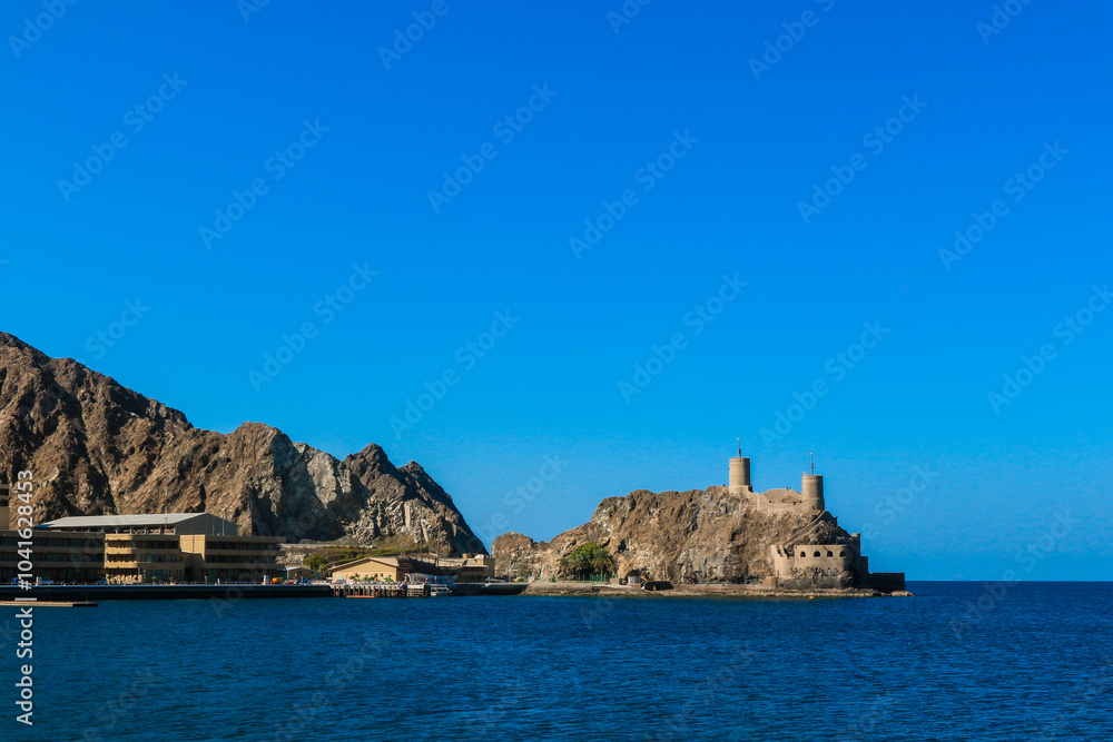 A picturesque view of the coastline in Muscat Oman featuring historic towers and rocky mountains under a clear blue sky A picturesque view of the coastline in Muscat Oman featuring historic towers and rocky mountains under a clear blue sky