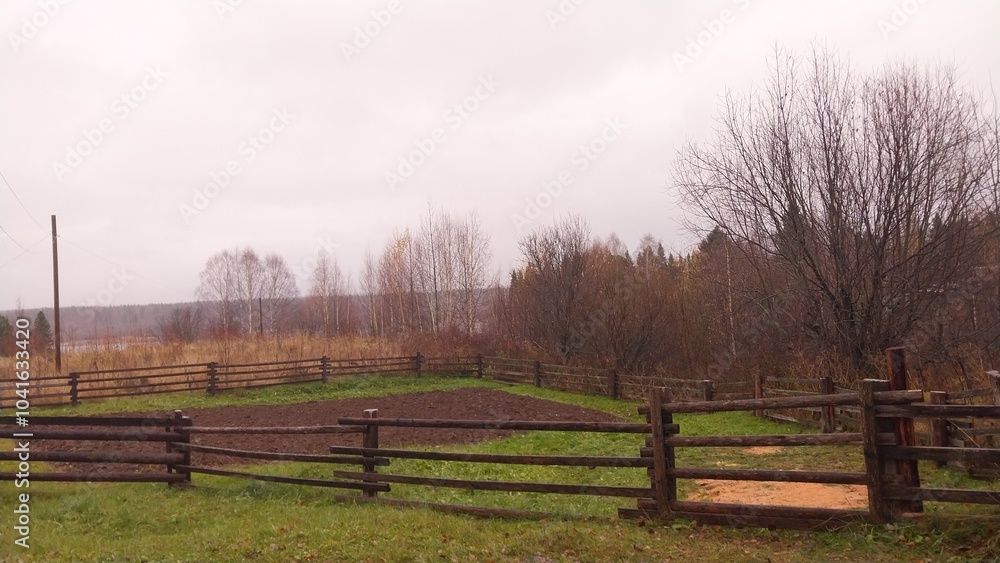 a rural landscape with a wooden fence in the foreground. The fence is made of horizontal wooden planks and encloses a grassy area. Beyond the fence, there is a field with bare trees a rural landscape with a wooden fence in the foreground. The fence is made of horizontal wooden planks and encloses a grassy area. Beyond the fence, there is a field with bare trees