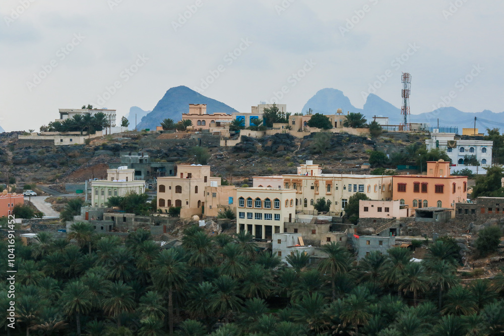 Naklejka premium A tranquil village in Oman nestled among hills and palm trees under a cloudy sky