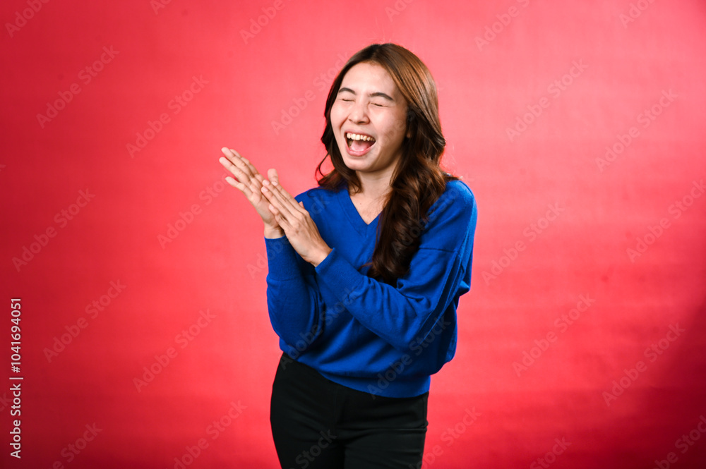 An Asian woman in a blue sweater, clapping her hands with a cheerful expression, standing against a red background. Her joyful energy and excitement are vividly displayed. An Asian woman in a blue sweater, clapping her hands with a cheerful expression, standing against a red background. Her joyful energy and excitement are vividly displayed.