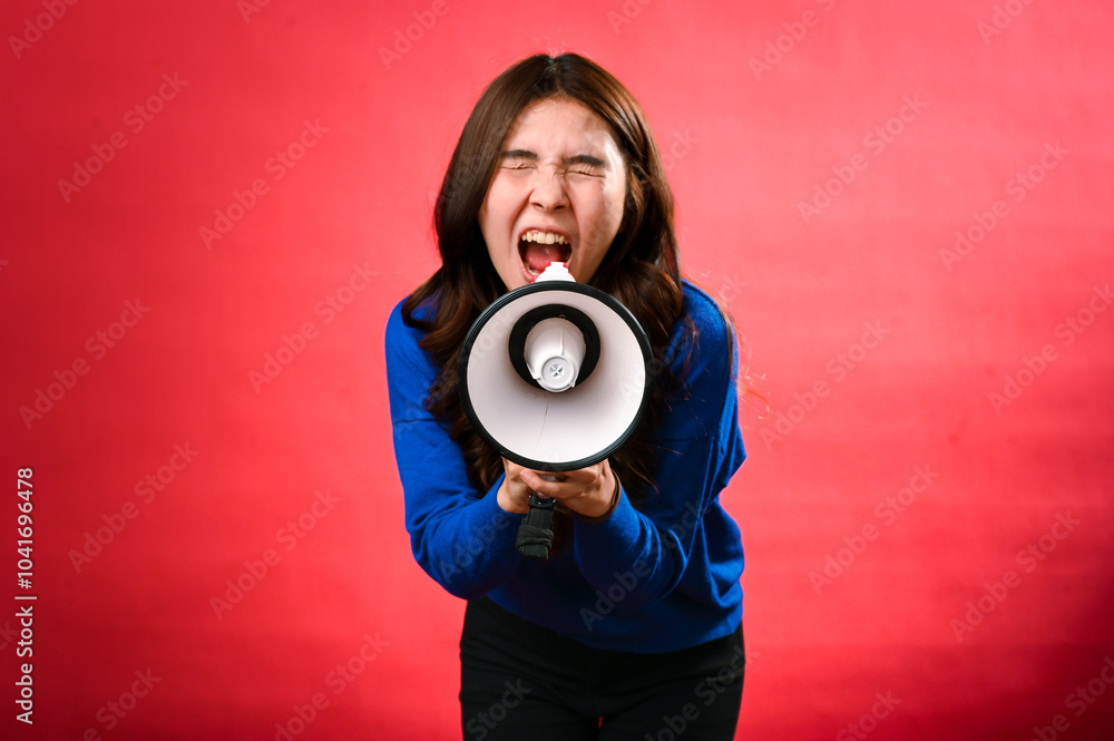 Fototapeta premium An Asian woman wearing a blue sweater is holding a red and white megaphone while speaking into it. She appears to be shouting or making an announcement. The background is solid red