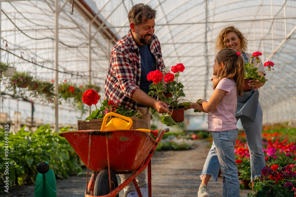 A family working together in a greenhouse, with the father placing flowers in a wheelbarrow, the daughter assisting, and the mother tending to plants. A family working together in a greenhouse, with the father placing flowers in a wheelbarrow, the daughter assisting, and the mother tending to plants.