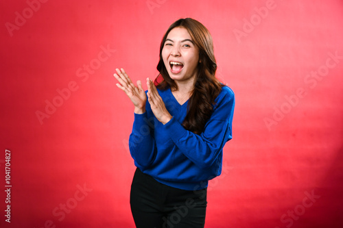 An Asian woman in a blue sweater, clapping her hands with a cheerful expression, standing against a red background. Her joyful energy and excitement are vividly displayed.