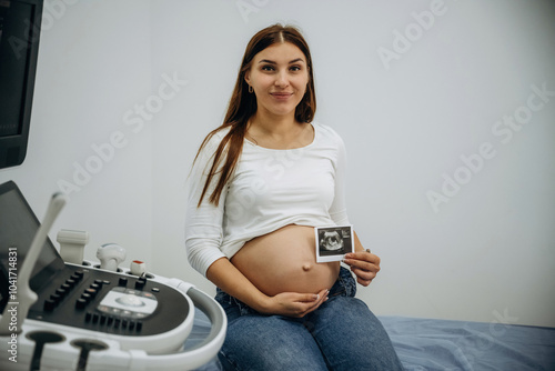 Pregnant woman is sitting in the clinic and holding image of ultrasonic test, conception of anticipation