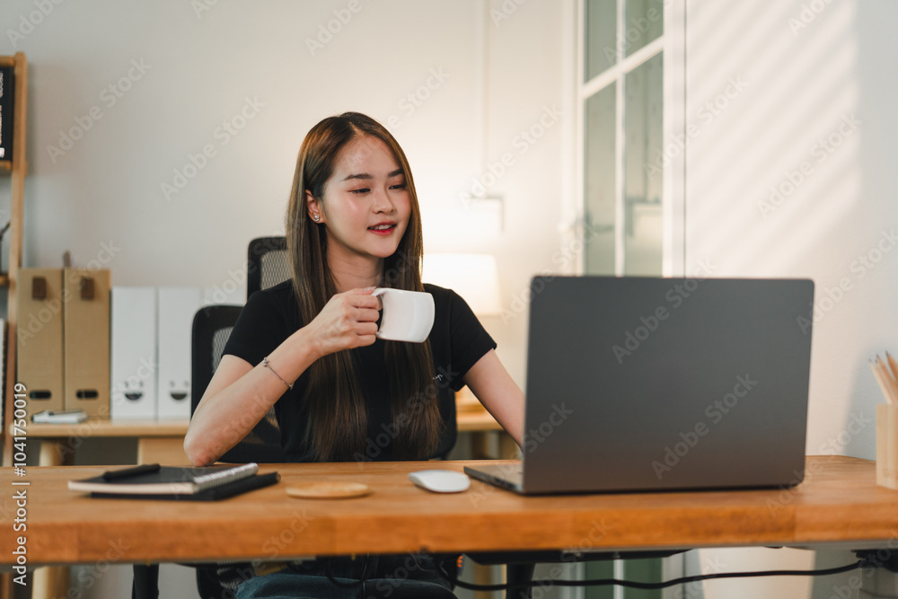 woman sitting at wooden desk, enjoying cup of coffee while working on laptop. modern workspace features clean design with natural light streaming in, creating productive atmosphere woman sitting at wooden desk, enjoying cup of coffee while working on laptop. modern workspace features clean design with natural light streaming in, creating productive atmosphere