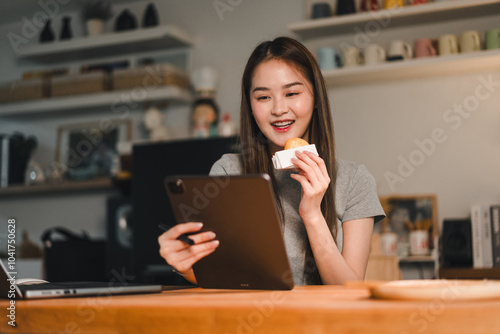 Enjoying snack while using tablet, young woman smiles as she engages with content on her device. cozy indoor setting features wooden table and shelves filled with various items, creating warm and