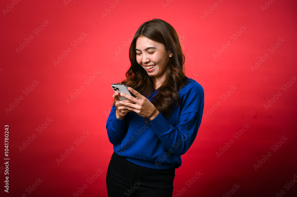 An Asian woman wearing a blue sweater is smiling as she looks down at her phone, engrossed in typing or reading a message. She stands against a bright red background in a cheerful, casual pose An Asian woman wearing a blue sweater is smiling as she looks down at her phone, engrossed in typing or reading a message. She stands against a bright red background in a cheerful, casual pose