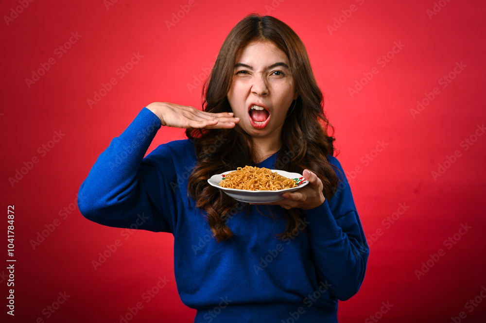 An Asian woman wearing a blue sweater holds a plate of noodles in one hand while gesturing excitement with the other hand near her mouth. She stands against a red background An Asian woman wearing a blue sweater holds a plate of noodles in one hand while gesturing excitement with the other hand near her mouth. She stands against a red background