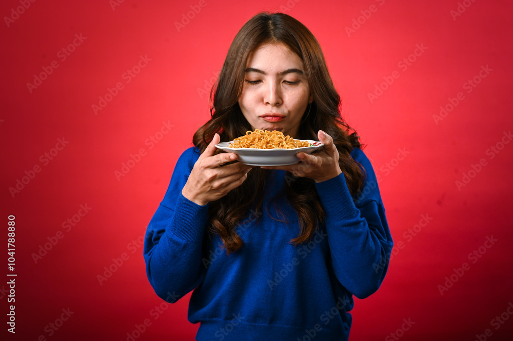 An Asian woman in a blue sweater holds a plate of noodles close to her face, licking her lips in anticipation. She stands against a red background with a playful and hungry expression An Asian woman in a blue sweater holds a plate of noodles close to her face, licking her lips in anticipation. She stands against a red background with a playful and hungry expression