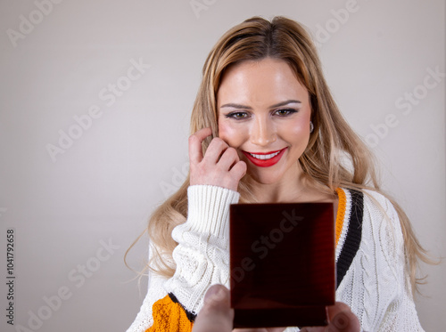 Photo of an attractive model with blonde hair smiling with her hand to the face while she is being asked to marry her with a man holding up a wedding ring box proposing or jewellery gift box photo