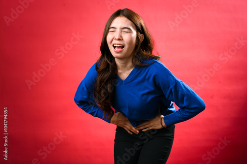 An Asian woman wearing a blue sweater bends forward with her hands clutching her abdomen, appearing to be in pain or discomfort. She winces with her eyes closed against a red background photo