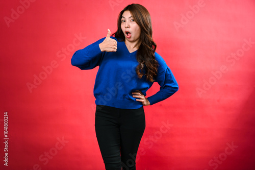 An Asian woman in a blue sweater and black pants stands confidently against a red background. She smiles while giving a thumbs-up gesture with her right hand and her left hand on her hip