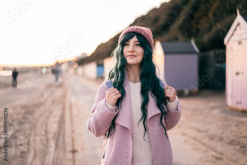 Stylish hipster woman with color hair in pink outfit and backpack walking along wooden beach huts on seaside. Off season Travel concept. Seasonal street fashion. Barbiecore style. Simple pleasures photo