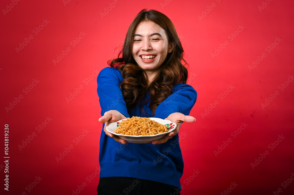 An Asian woman wearing a blue sweater is enthusiastically holding out a plate of noodles toward the camera. She stands against a red background, expressing excitement and energy in her pose An Asian woman wearing a blue sweater is enthusiastically holding out a plate of noodles toward the camera. She stands against a red background, expressing excitement and energy in her pose
