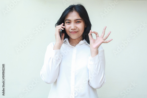 pleased satisfied asian business woman showing ok signs while speaking on smatphone wearing white formal suit shirt, approval good gesture, okay symbols, standing isolated looking at camera